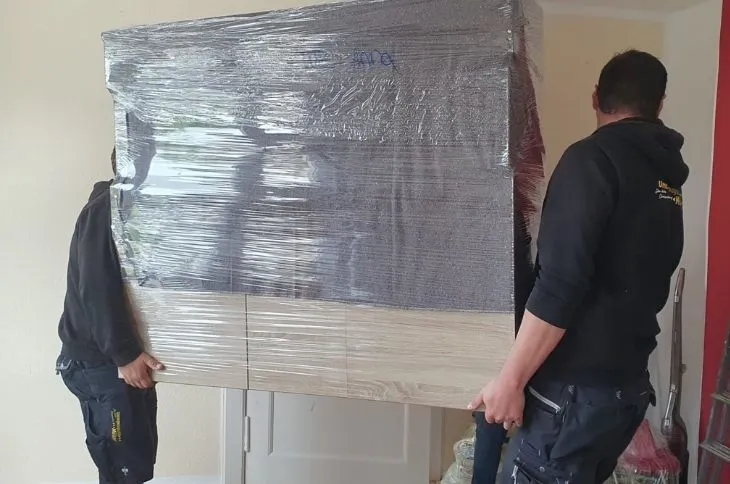 zwei umzugshelfer tragen grossen verpackten schrank im raum